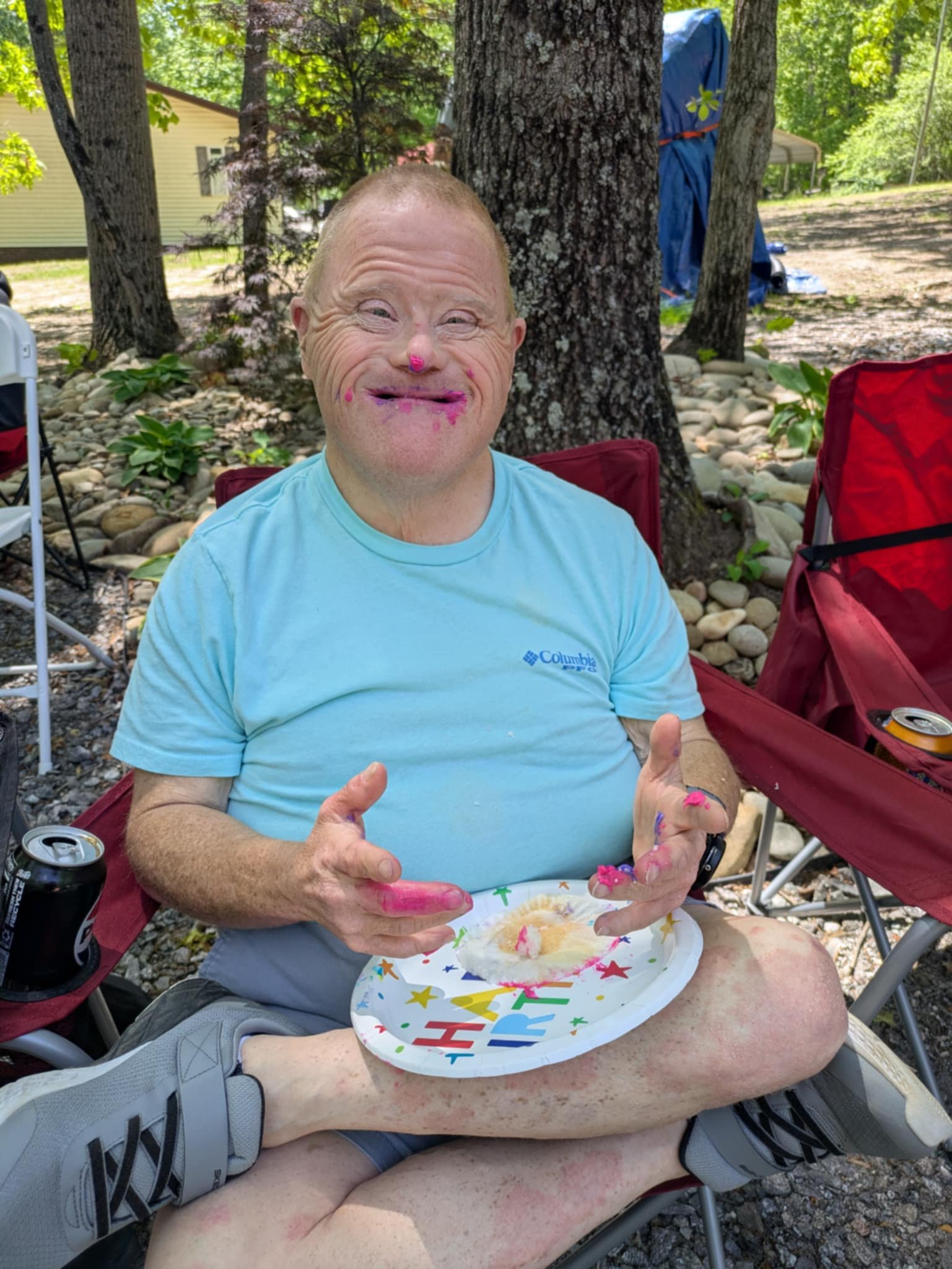 A man with messy pink face and hands, smiling, sitting outdoors at a party, holding a paper plate with cake, and wearing a light blue Columbia t-shirt. There are trees, rocks, and chairs in the background.