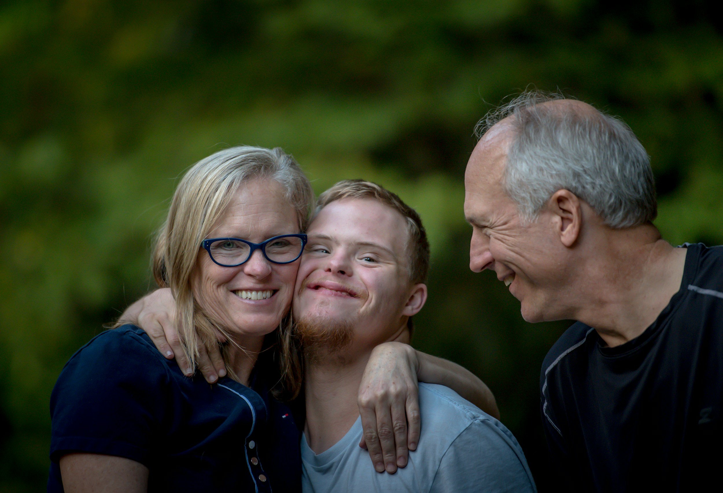 Three happy people, an older woman with glasses, a young man, and an older man, are smiling and embracing outdoors with a blurred green background.