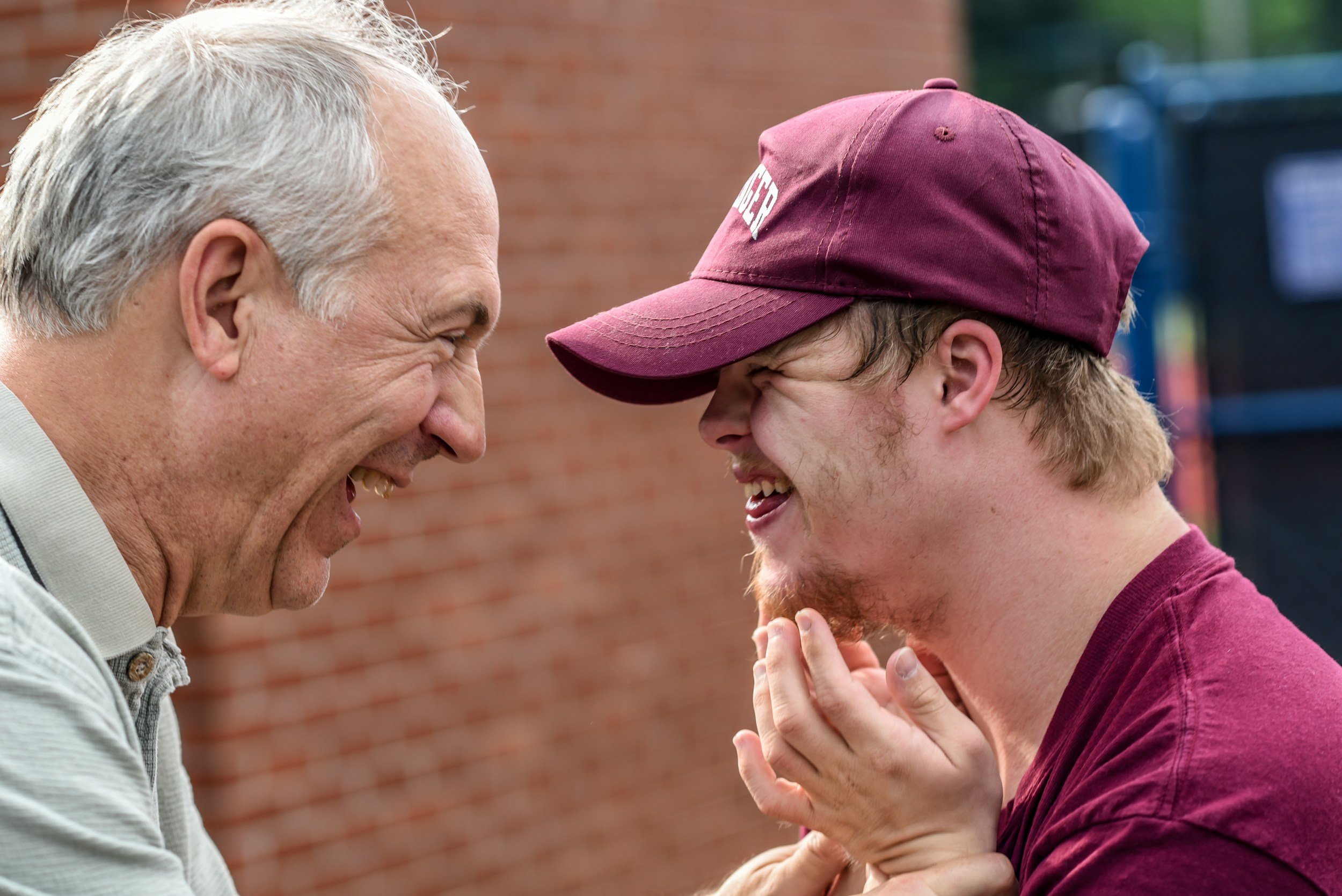 Two men playing a game of ring toss outdoors, with one wearing a grey shirt and the other wearing a maroon baseball cap and maroon shirt, both laughing.