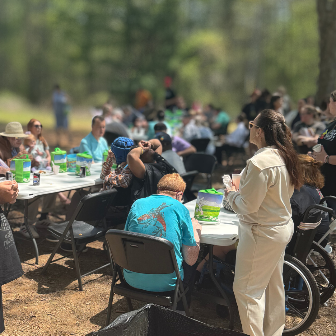 People gathered outdoors for a meal or celebration at a long table, with some in wheelchairs and wearing casual clothing, in a wooded area.