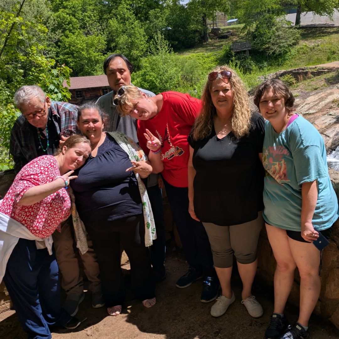 Group of eight people posing outdoors near rocks and greenery on a sunny day.