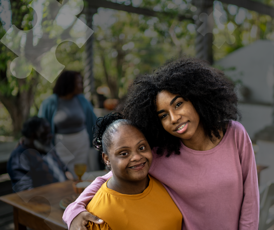 Two women smiling and hugging outdoors during the daytime, with others in the background at a gathering or social event.