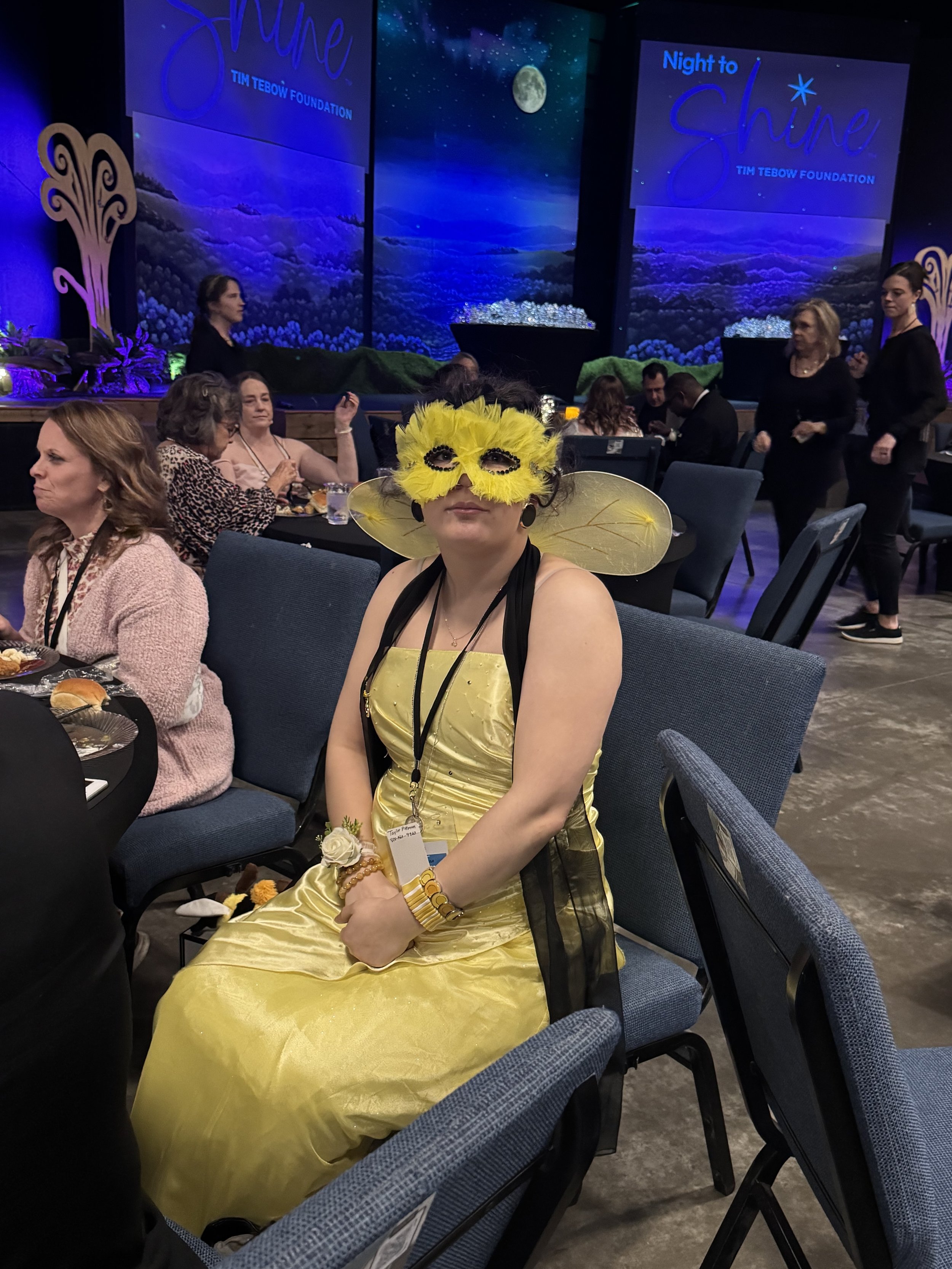 Woman dressed as a bee with a yellow dress, yellow wings, and a yellow feather mask sitting at a table at a formal event or conference. The background features a stage with a large screen displaying the words 'Night to Shine' and 'Tim Tebow Foundation' with a moon and starry sky imagery, and several other people are seated or walking around.