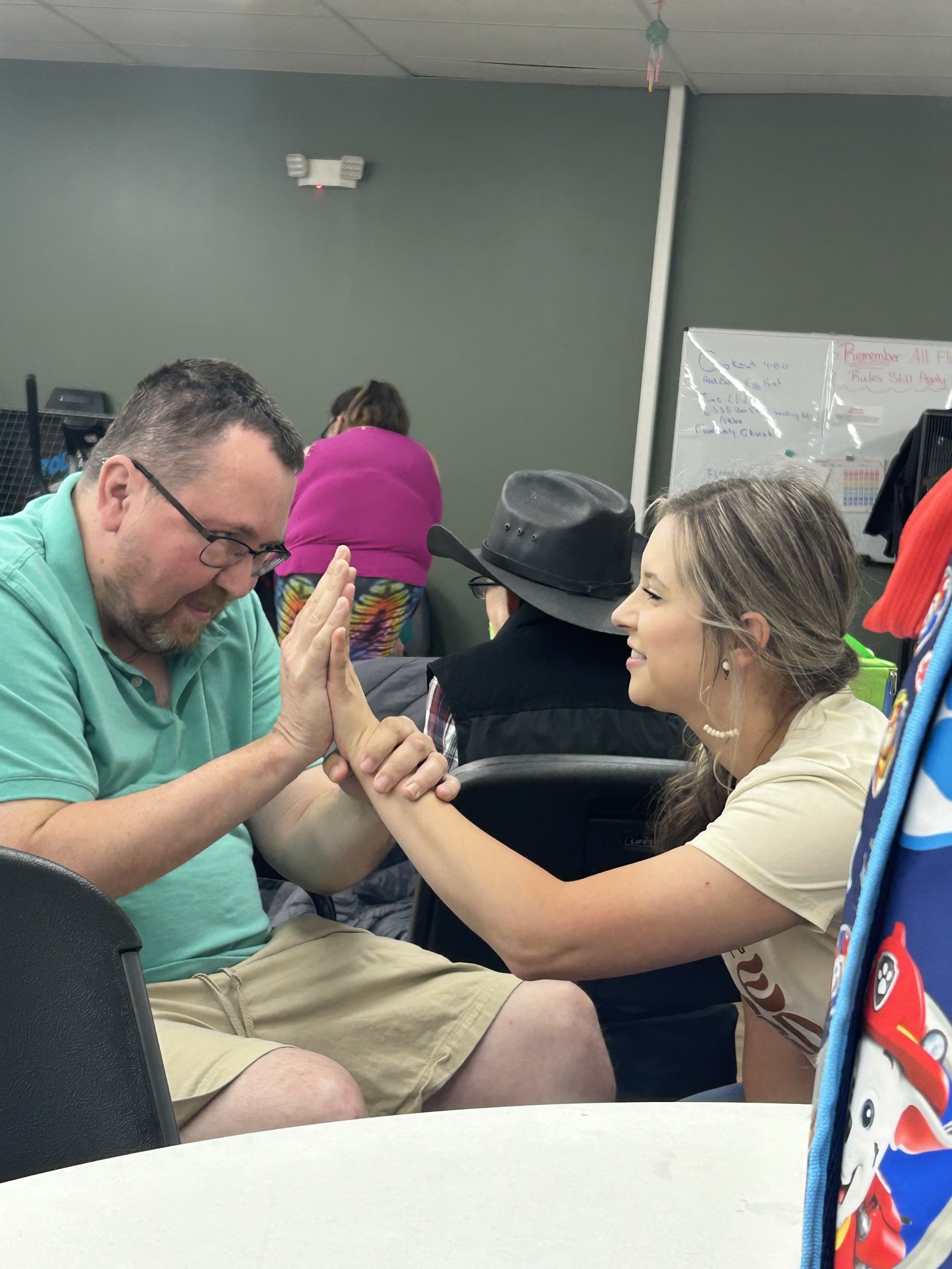 A man and woman engaging in a friendly arm wrestling match at a table in a classroom or community center, smiling and enjoying the moment.