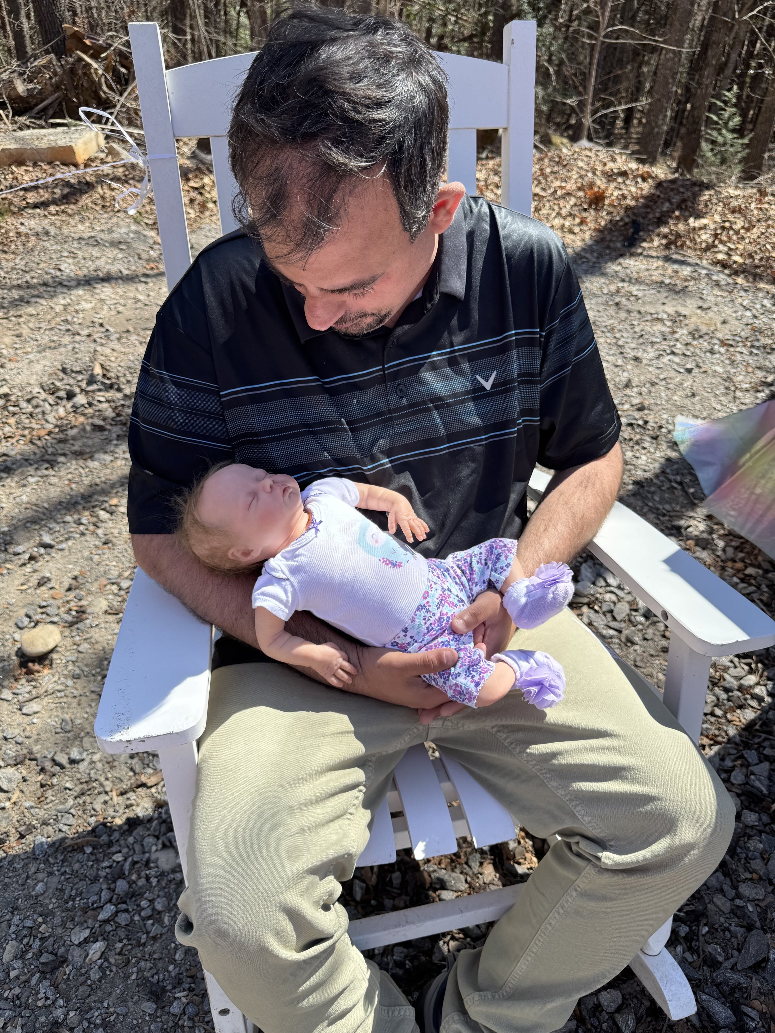 A man sitting outdoors on a white wooden chair, holding a newborn baby on his lap. The man is looking down at the baby, who is wearing a white shirt, floral pants, and purple booties. The background shows a gravel ground and trees.
