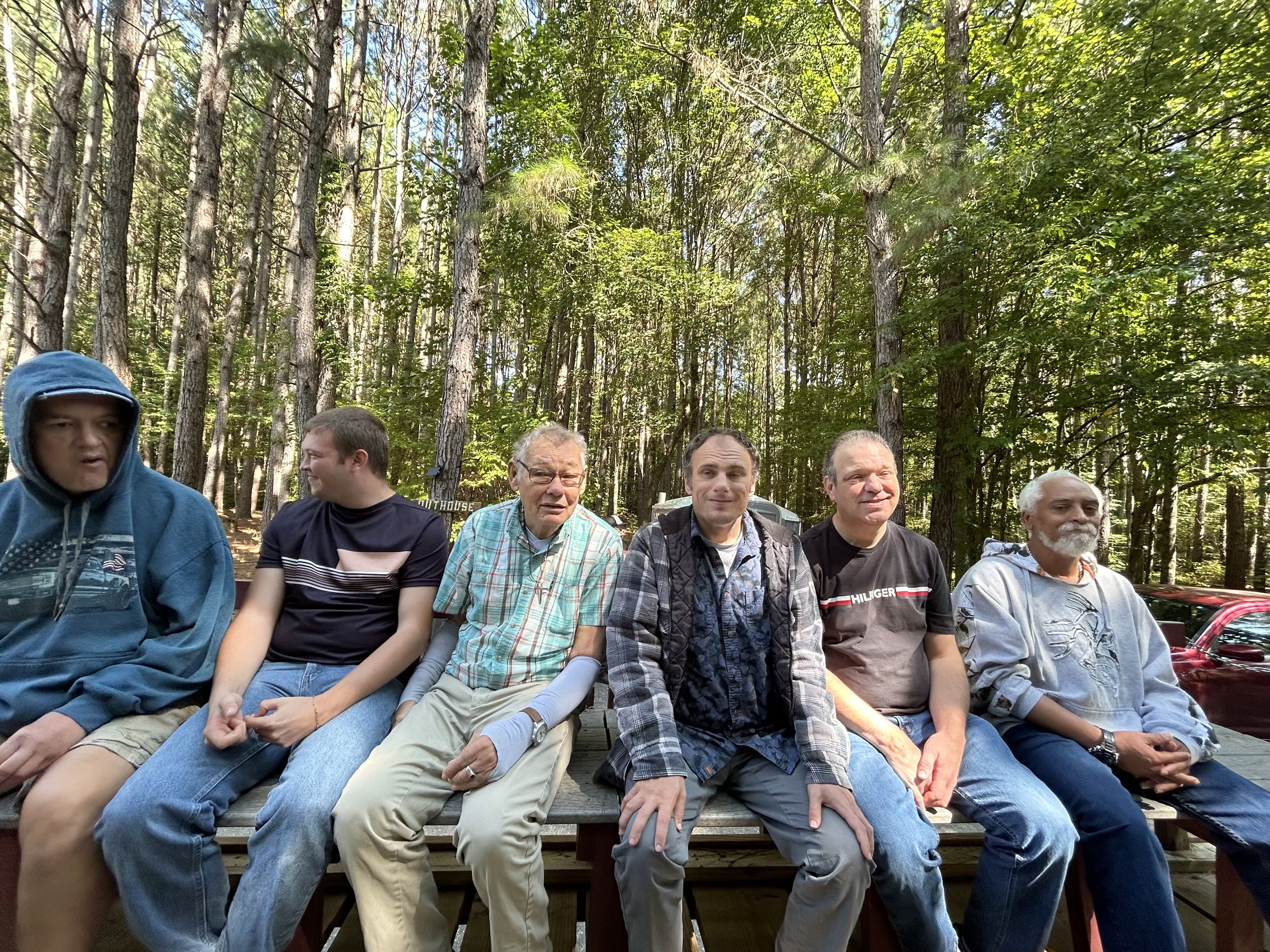 Six men sitting on a bench outdoors in a wooded area with green trees.